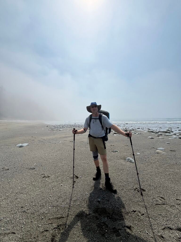 A tall hiker carrying a heavy pack uses two aluminum trekking poles to balance on the West Coast Trail.